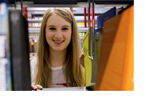 Eine junge Studentin schaut durch ein Bücherregal. (Foto: Julien Fertl)